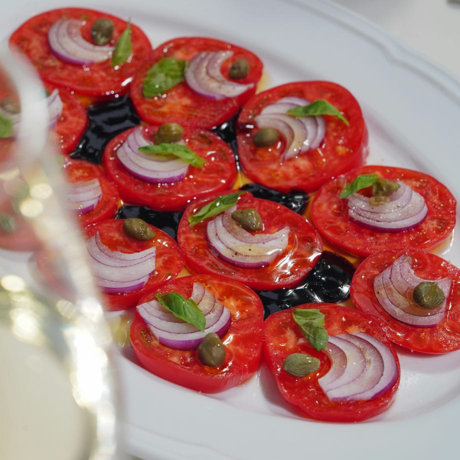 Frischer Tomaten-Caprese-Salat mit roten Zwiebeln, Basilikum und Kapern, beträufelt mit Balsamico-Glasur auf einem weißen Teller.